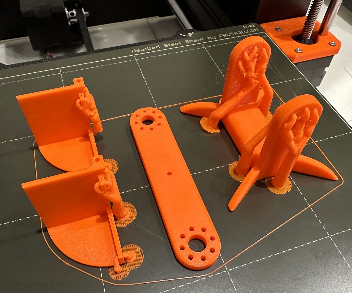 Freshly 3D-printed orange parts on a printer bed: two motor brackets, a flat leg bar, and a foot.